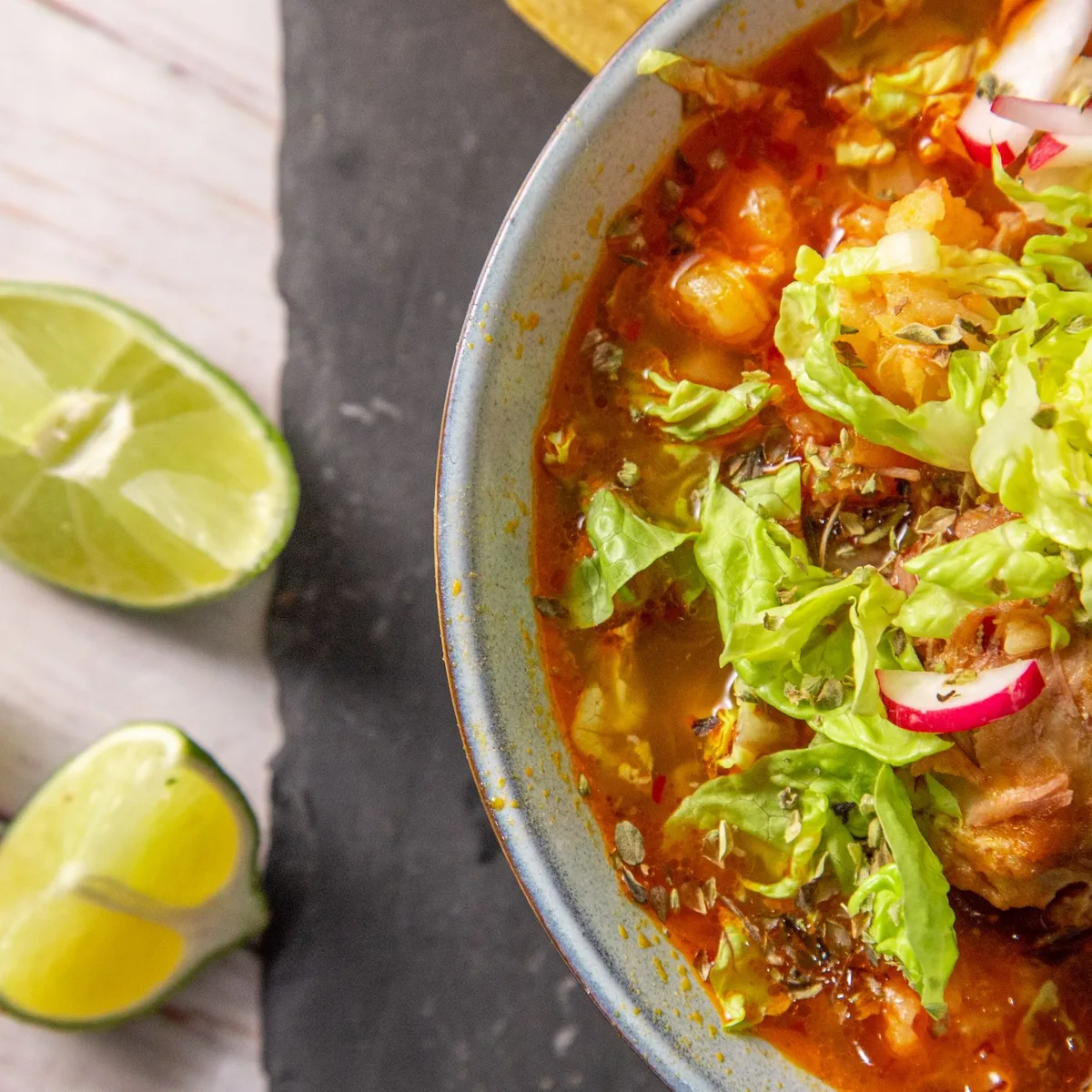 A bowl of red Mexican pozole topped with shredded lettuce, radish and oregano, limes alongside