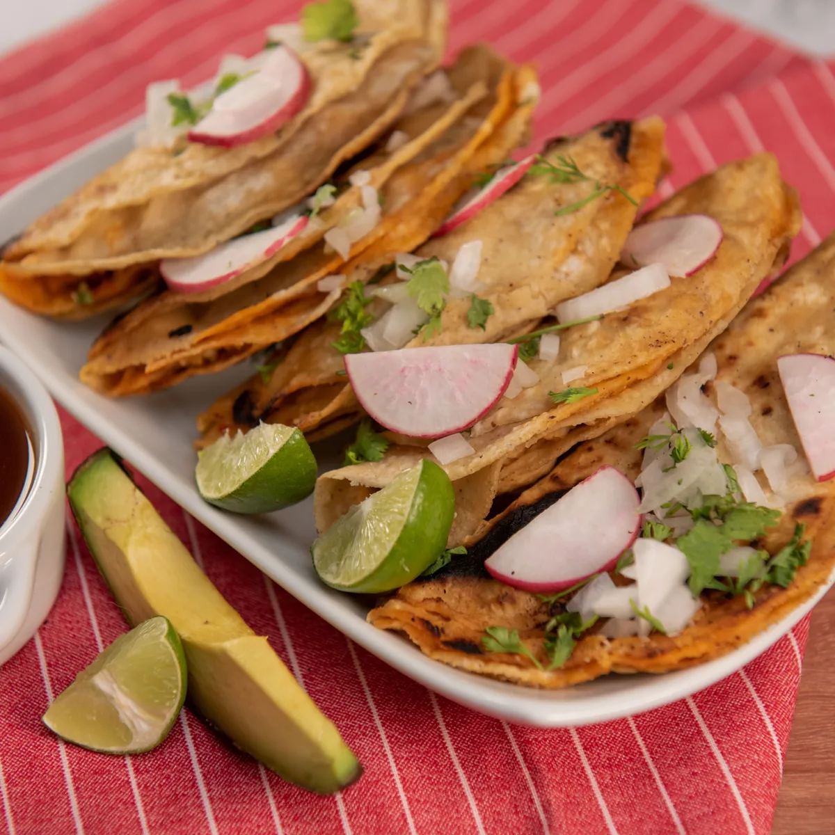 Griddled birria tacos topped with radish, onion and cilantro beside a cup of consome for dipping