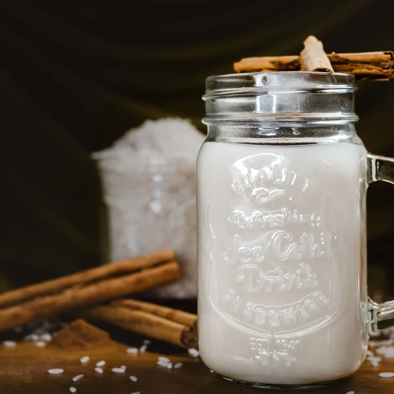 A glass jar of pale horchata with a cinnamon stick, beside raw rice and cinnamon