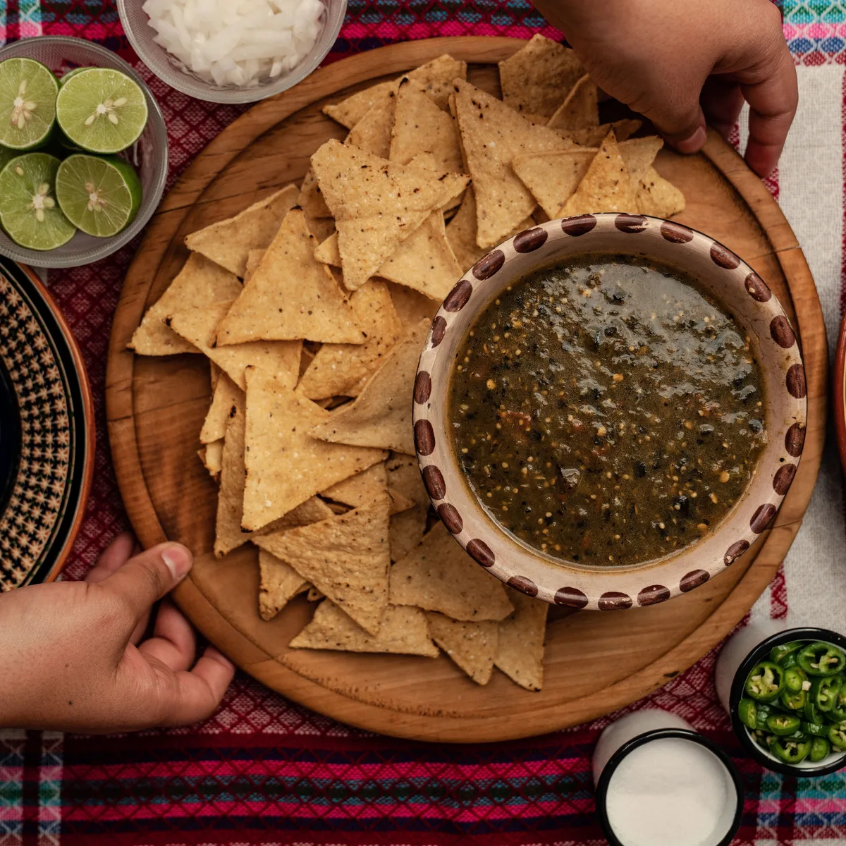A clay bowl of roasted tomatillo salsa verde on a board with tortilla chips, limes and sliced jalapenos