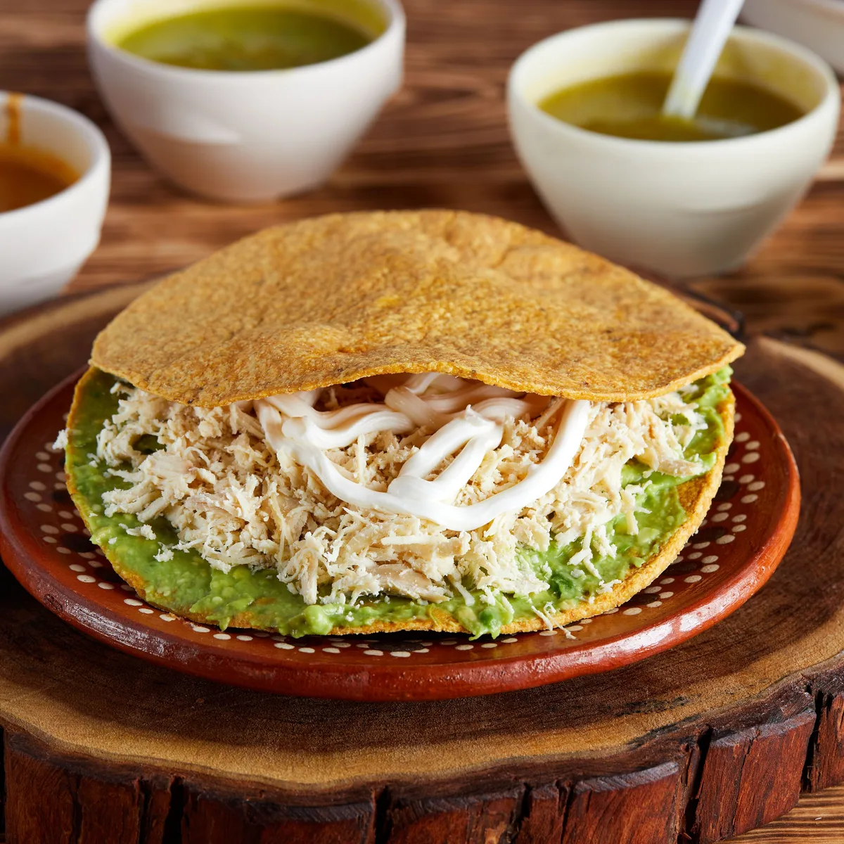 A crisp tostada topped with shredded chicken, avocado and crema, with bowls of salsa behind